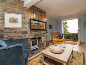 A cozy living room with a stone fireplace, a blue sofa, a yellow armchair, and a large window with a view of greenery. - Rookery Cottage, Lynton (Lynton)
