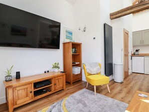 A living room with a TV, wooden entertainment unit, a yellow chair, and a kitchen area. - Harvenna Cottage, Crackington Haven (Bude)