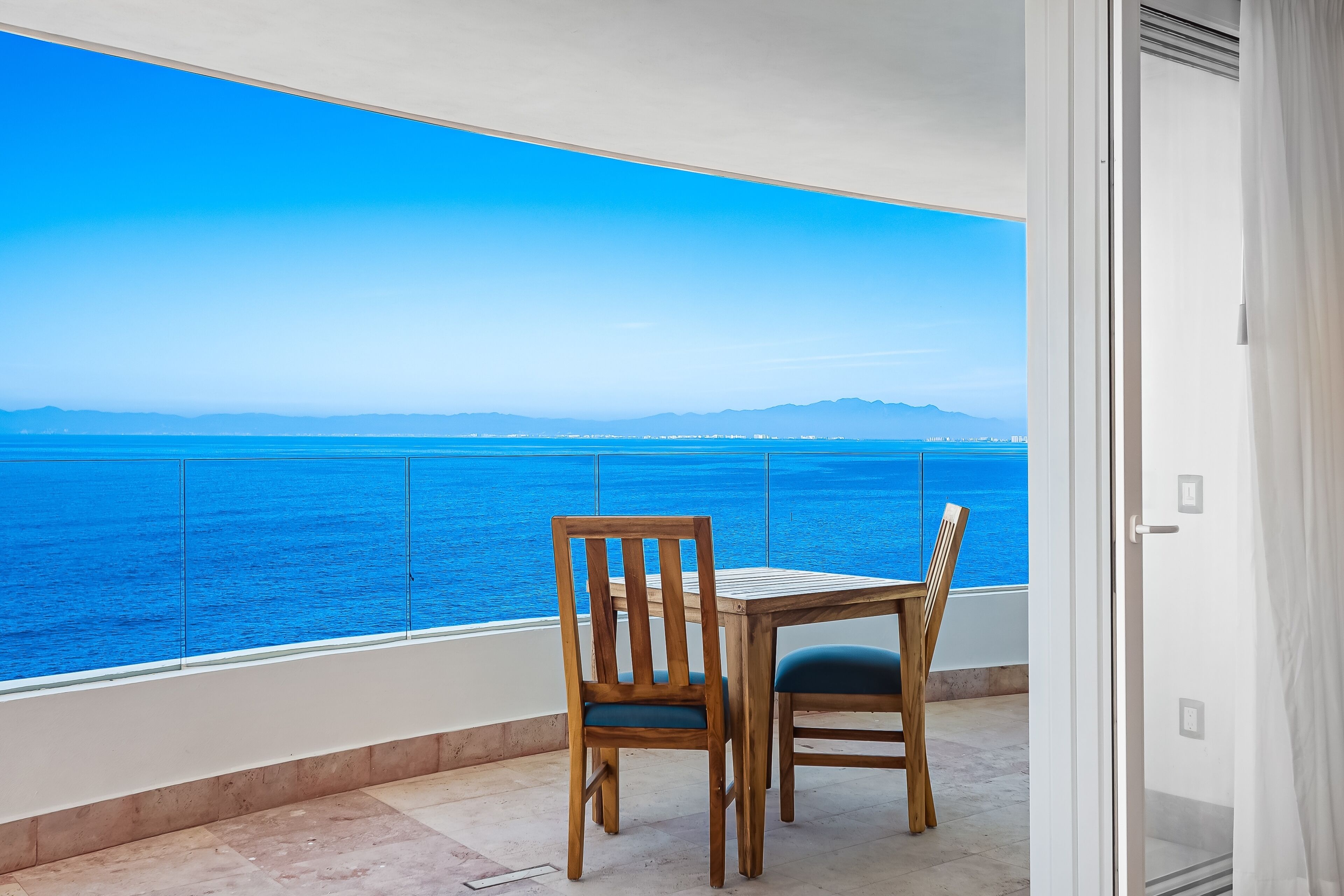 A balcony with a table and chairs, overlooking a body of water and distant mountains.