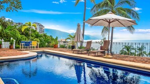 A swimming pool with lounge chairs and umbrellas, surrounded by palm trees and a view of the ocean. - Ocean-view Conchas Chinas 3BR near beaches, dining, nightlife (Puerto Vallarta)