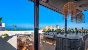 An outdoor terrace with a view of the ocean, featuring wicker furniture, a sofa, and a bar area.