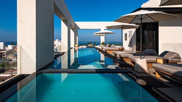 A rooftop infinity pool with lounge chairs and umbrellas, overlooking a cityscape and the sea.