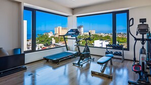 A modern gym with a treadmill, bench, dumbbells, and exercise equipment, featuring large windows with a city view.