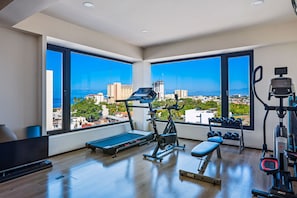 A modern gym with a treadmill, bench, dumbbells, and exercise equipment, featuring large windows with a city view. - 2BR/2BA 5 De Diciembre condo in Puerto Vallarta (Puerto Vallarta)