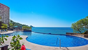 Ein Pool mit geschwungener Form, umgeben von Liegestühlen und Blick aufs Meer.
