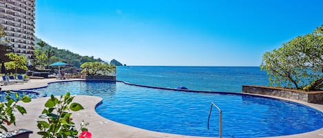 A swimming pool with a curved design, surrounded by lounge chairs and a view of the sea.