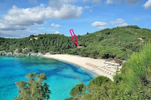 Un paysage côtier avec une plage, une eau d’un bleu limpide et des collines en arrière-plan.