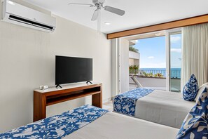 Ein Hotelzimmer mit Flachbildfernseher auf einem Holzboden, zwei Betten mit blau-blumigen Überwürfen, einem Deckenventilator und einem Balkon mit Meerblick.