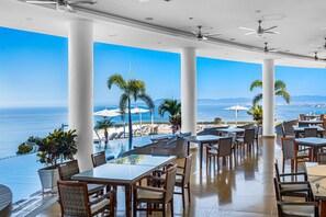 Un vaste espace extérieur aménagé pour les repas, avec des tables et des chaises, offrant une vue sur une piscine et l’océan.