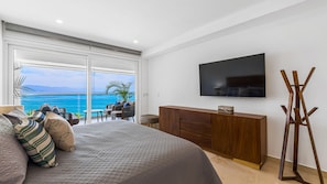 Une chambre à coucher avec un grand lit, une télévision, une commode en bois et un balcon avec vue sur la mer.