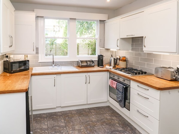 Une cuisine moderne avec des meubles blancs, un plan de travail en bois, une cuisinière, un four à micro-ondes et une fenêtre avec des stores.