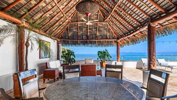 A covered outdoor seating area with a round table and chairs, a thatched roof, and a view of the ocean. - Elegant multi-level penthouse with rooftop Jacuzzi, bay views, pool & game room (Puerto Vallarta)