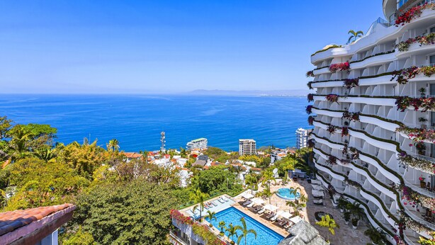 A high-rise hotel with multiple balconies adorned with flowers, a swimming pool, and a view of the ocean. - Elegant bayview suite with terrace access, full kitchen, pool, hot tub & gym (Puerto Vallarta)