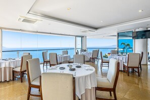 A dining area with round tables, chairs, and a large window overlooking the sea. - Elegant hillside villa with panoramic views, pool, hot tub & luxe interiors (Puerto Vallarta)