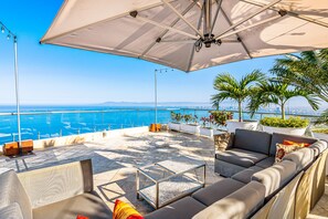 A terrace with a sofa, a coffee table, and an umbrella, overlooking a coastal view. - Elegant hillside villa with panoramic views, pool, hot tub & luxe interiors (Puerto Vallarta)