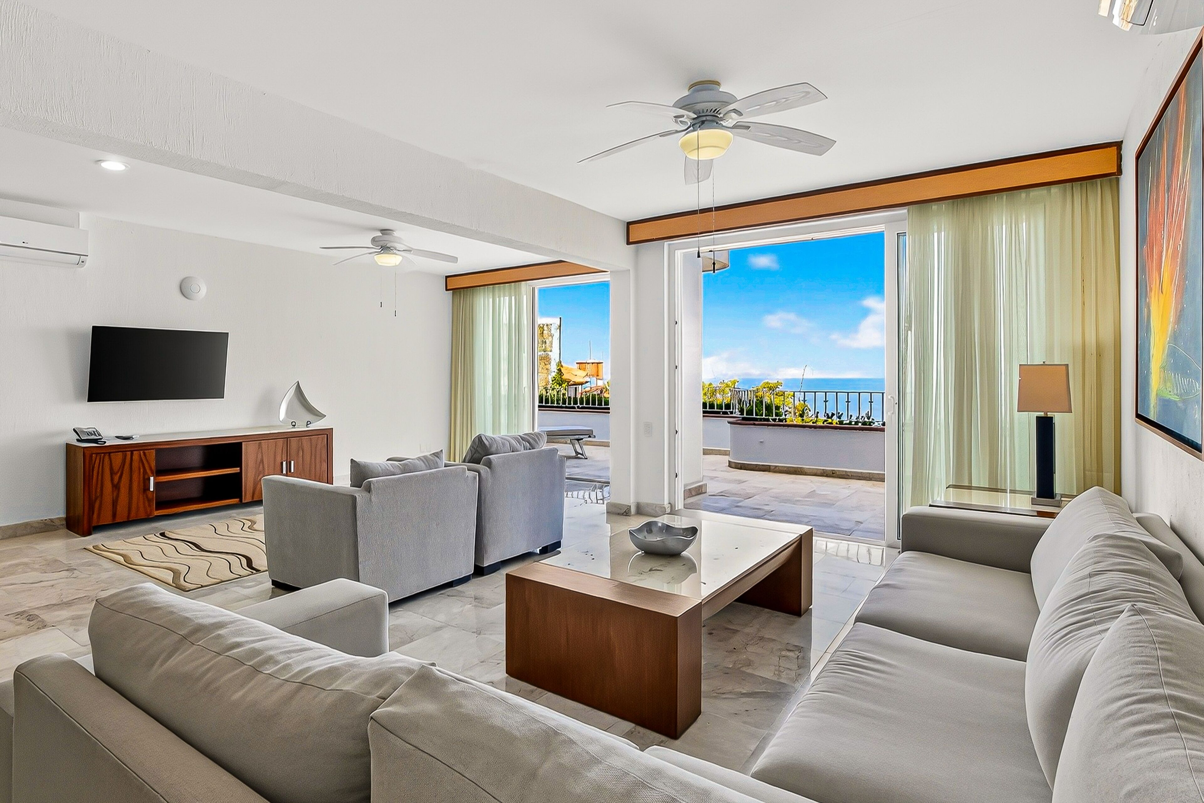 A modern living room with a flat-screen TV, sofas, a coffee table, and a balcony with a view.