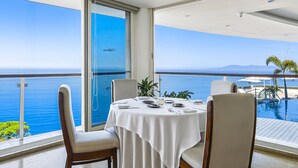 A dining area with a white tablecloth, four chairs, and a view of the sea through large windows. - Stunning oceanview suite with cliffside infinity pool, terrace & gourmet kitchen (Puerto Vallarta)