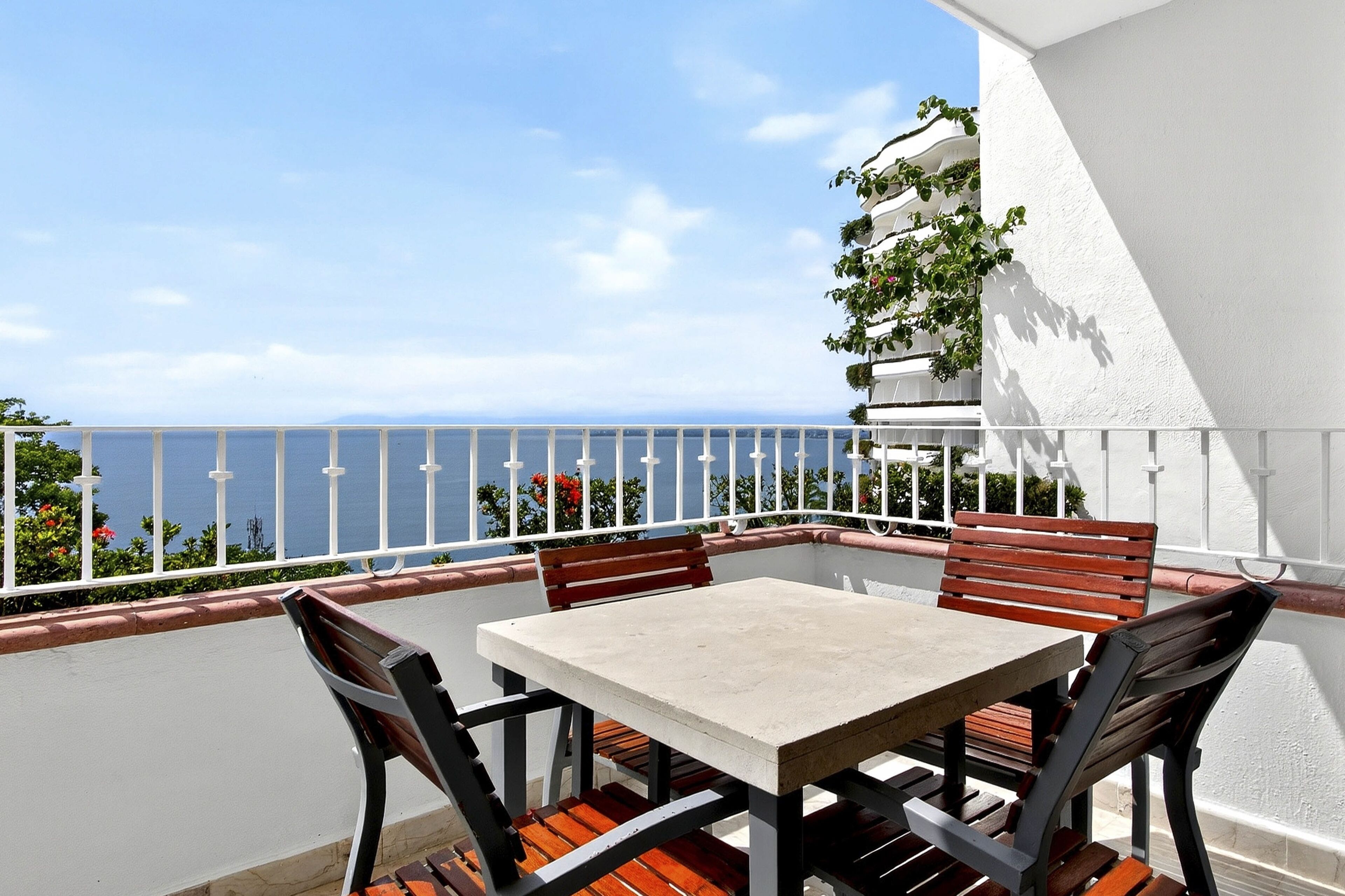 En balkon med et bord og stole, et hvidt gelænder og udsigt over havet.