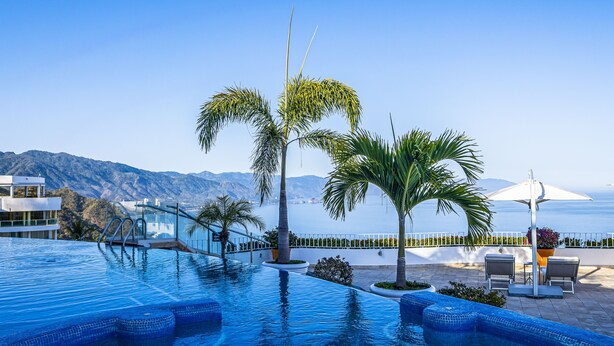 A swimming pool with a view of mountains and the sea, surrounded by palm trees and a lounge area with chairs and a table. - Stunning oceanview suite with cliffside infinity pool, terrace & gourmet kitchen (Puerto Vallarta)