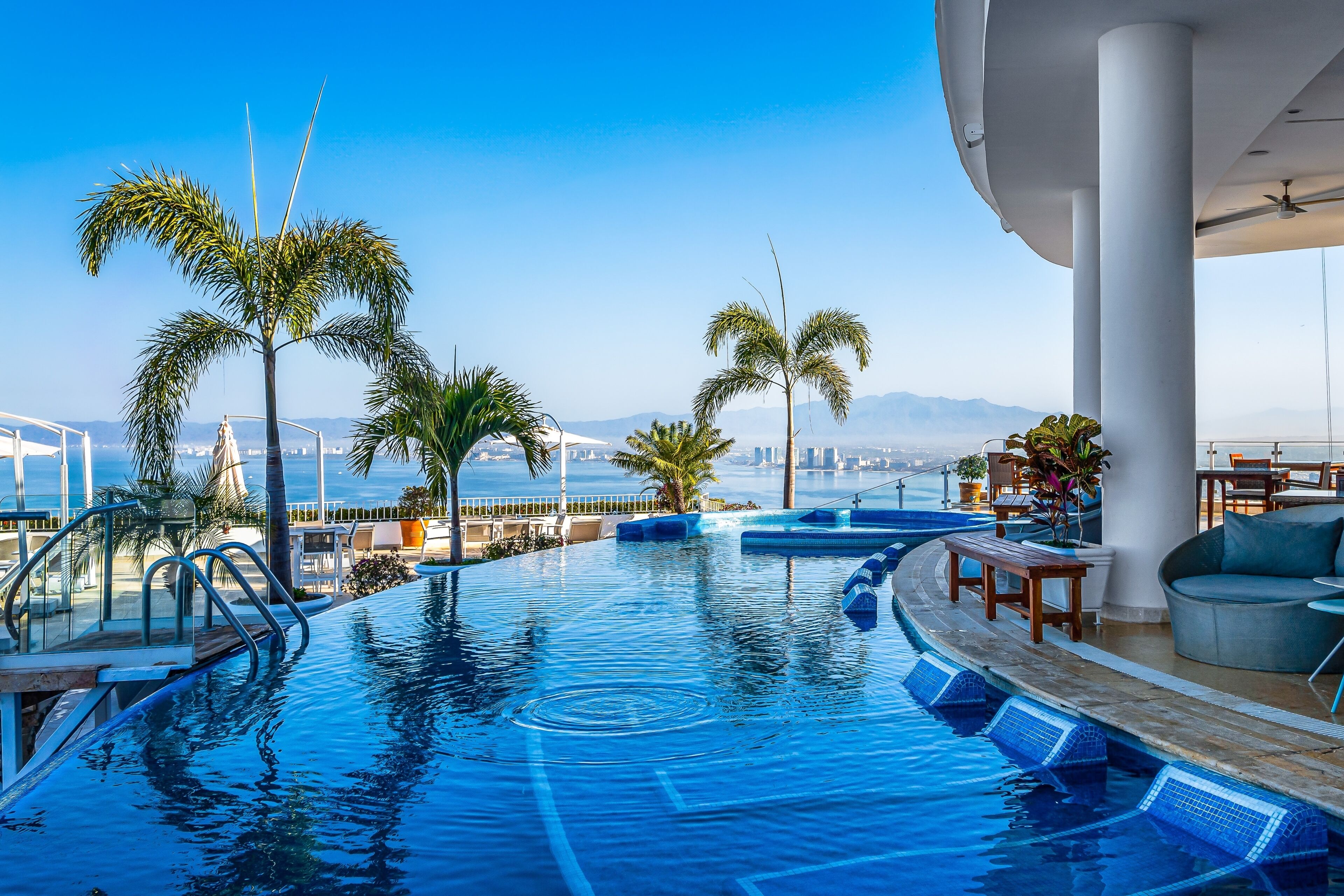 A rooftop infinity pool with palm trees, a view of the city, and a dining area with outdoor seating.
