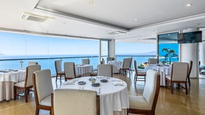 Un restaurante con mesas redondas dispuestas para comer, amplias ventanas que dan al mar y un televisor de pantalla plana montado en la pared.