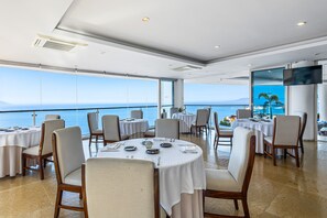 Ein Restaurant mit runden Tischen, großen Fenstern mit Meerblick und einem an der Wand befestigten Flachbildfernseher.