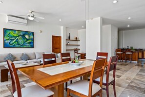 Un vaste espace de vie comprenant une table à manger en bois, des sièges confortables, un ventilateur de plafond moderne et une peinture abstraite aux couleurs vives.