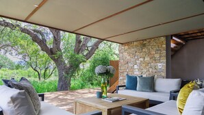 An outdoor seating area with a stone wall, wooden furniture, and a view of trees. - Villa Nsovo (Hoedspruit)