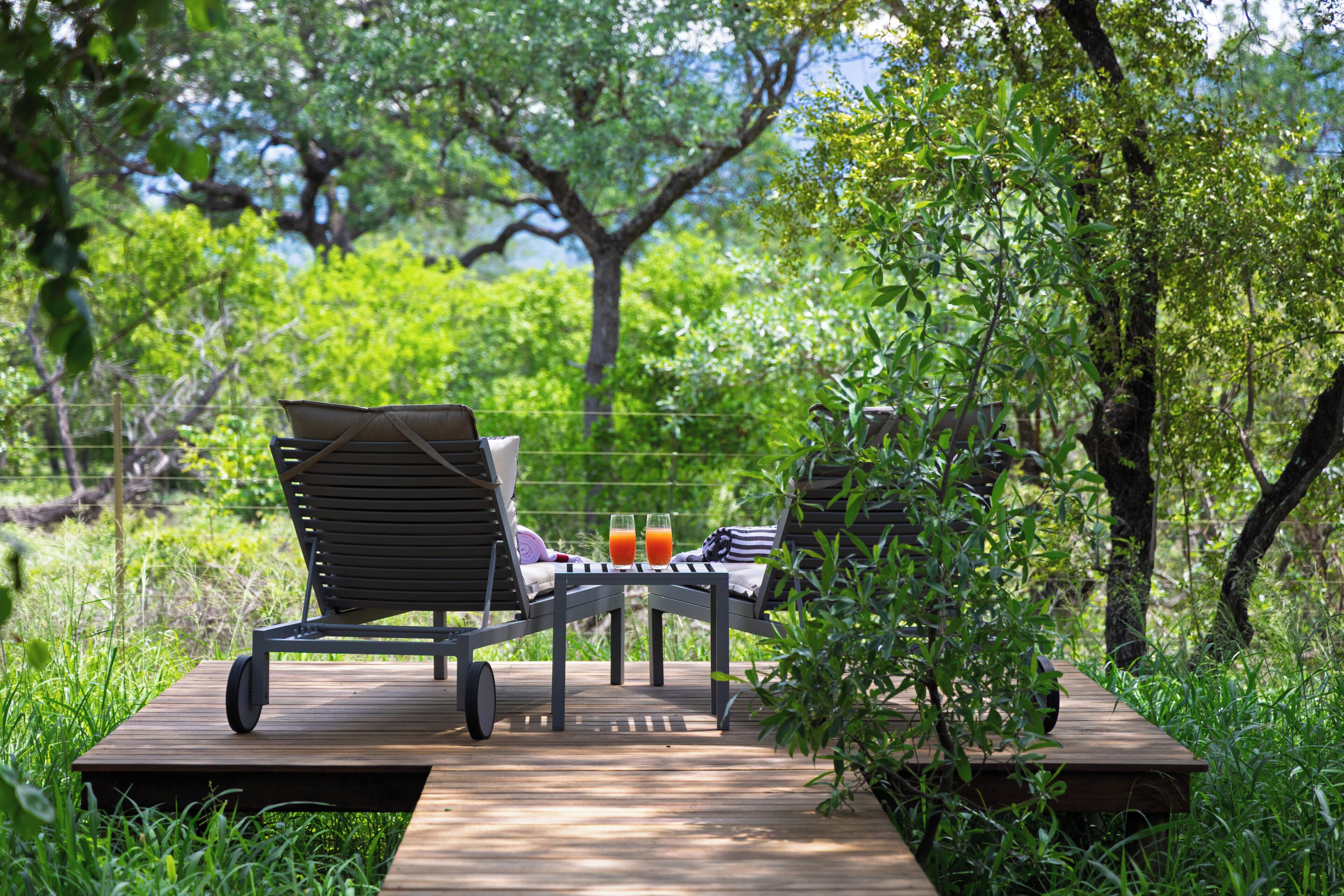 A wooden deck with two lounge chairs and a table, surrounded by greenery and trees.