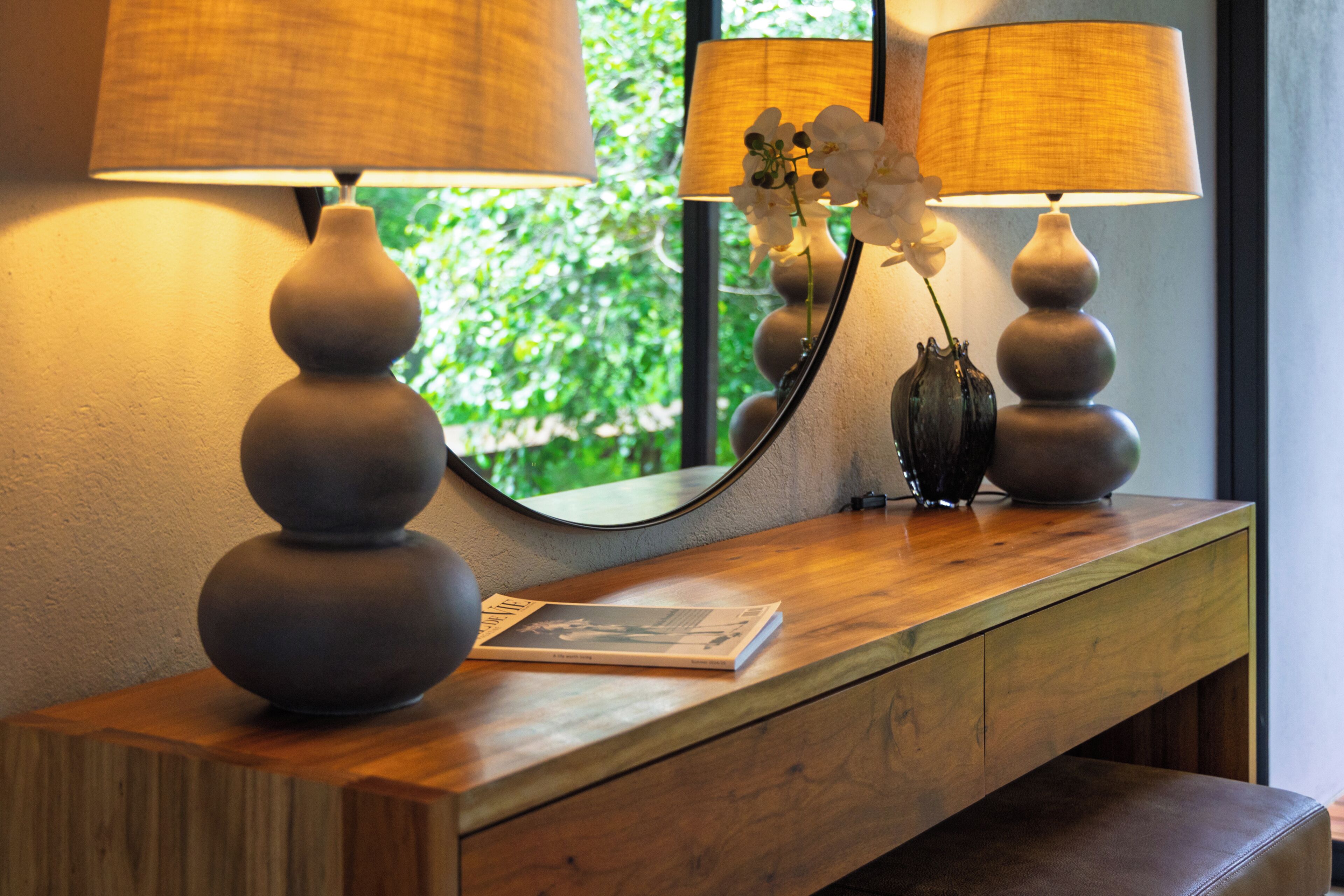 A wooden console table with two lamps, a vase with flowers, and a mirror reflecting greenery.