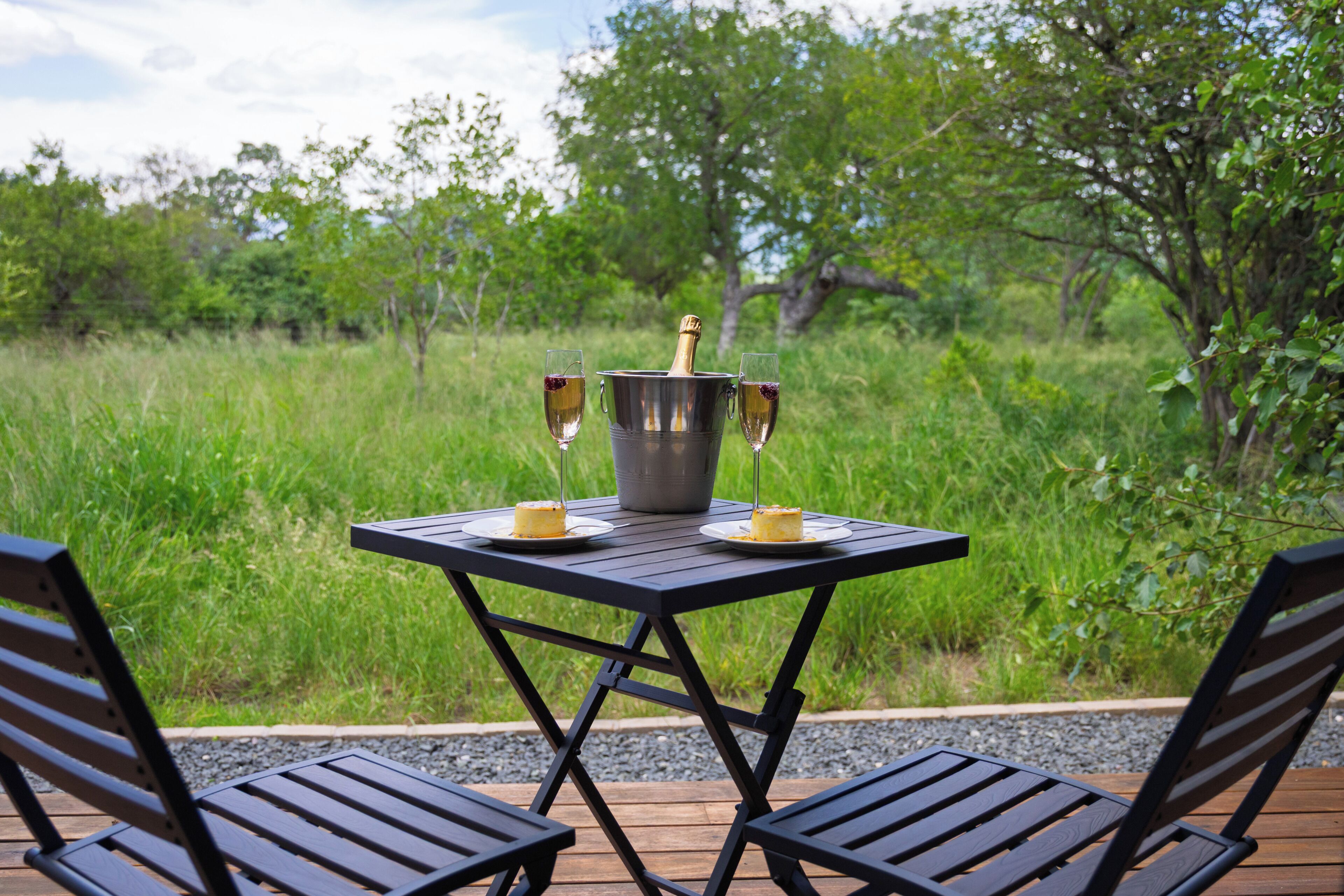 A table with a bottle and glasses, surrounded by chairs, set on a deck with a grassy background.