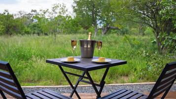 A table with a bottle and glasses, surrounded by chairs, set on a deck with a grassy background.