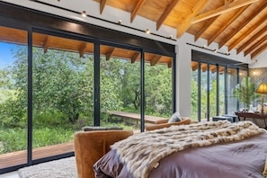 A bedroom with large windows, a wooden ceiling, and a fur throw on the bed. - Villa Nsovo (Hoedspruit)