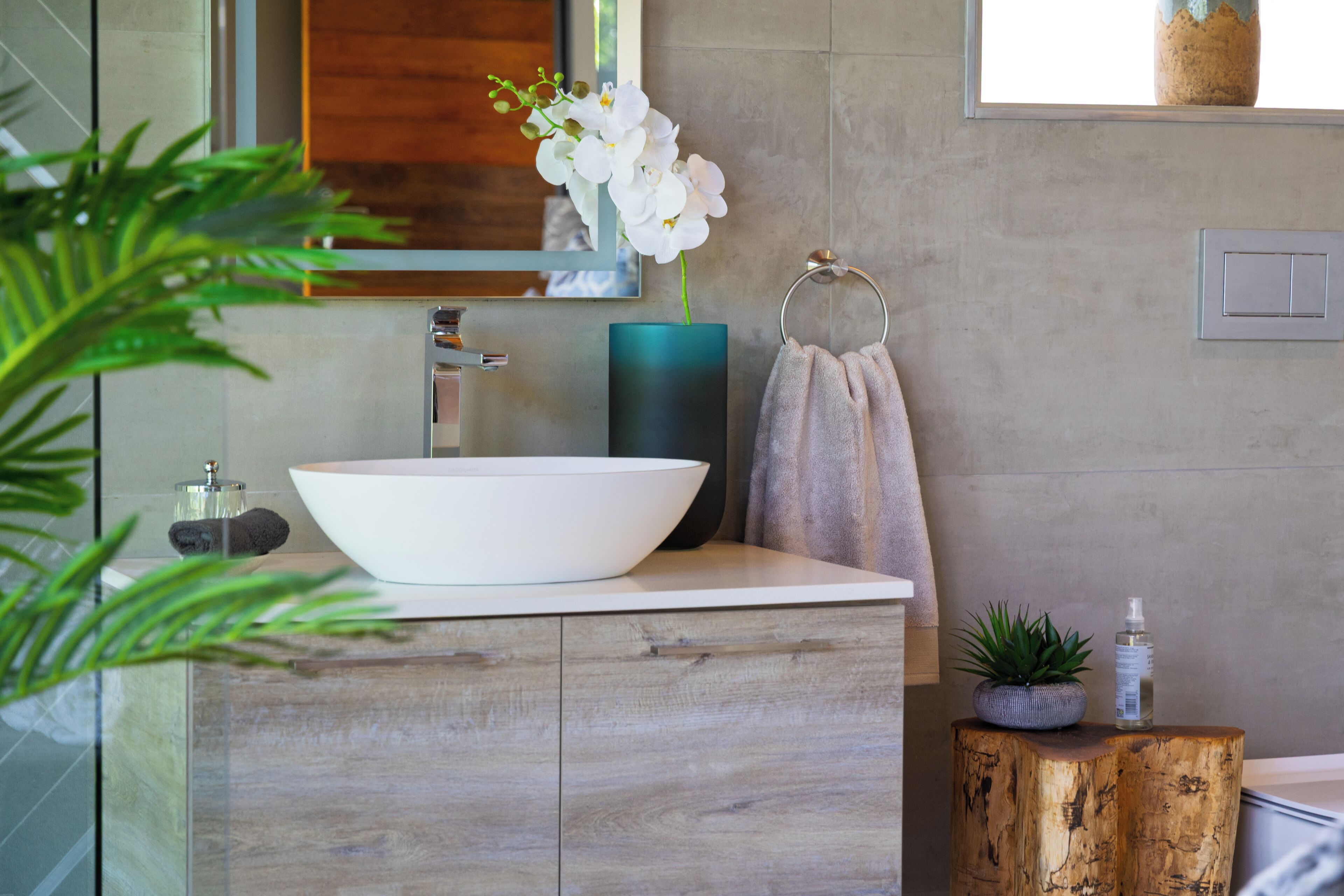 A bathroom with a large white sink, a mirror, a towel ring, a potted plant, and a wooden stool.