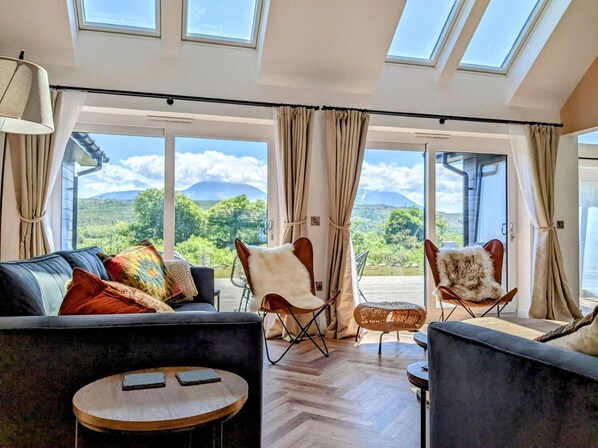 A spacious room with a large sofa, two armchairs, a small round table, and a view of a mountainous landscape through large windows.
