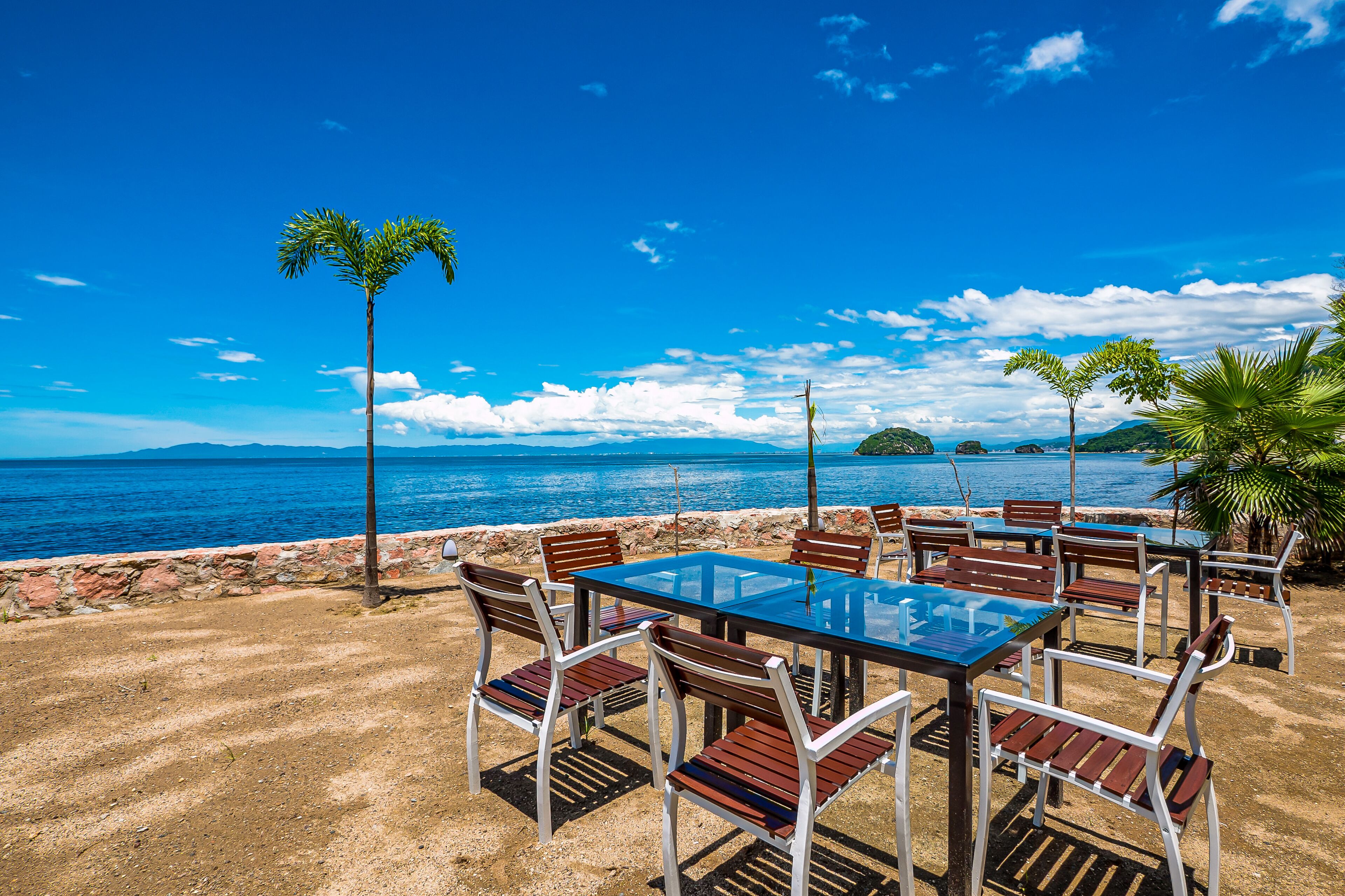 An outdoor seating area with a glass table and chairs, overlooking a body of water and distant land.