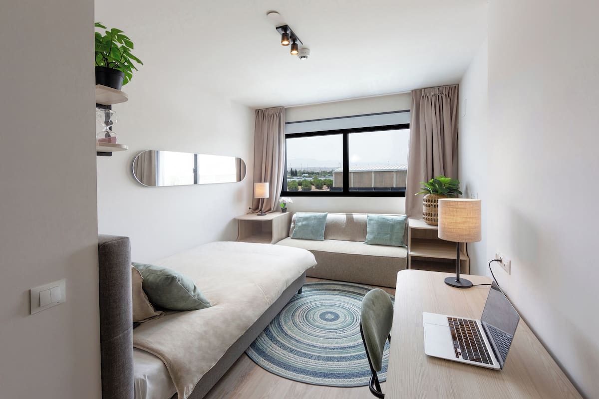 Ein modernes Hotelzimmer mit einem Bett, einem Schreibtisch mit Laptop, einem Fenster mit Vorhängen, einer Pflanze und einem Spiegel an der Wand.
