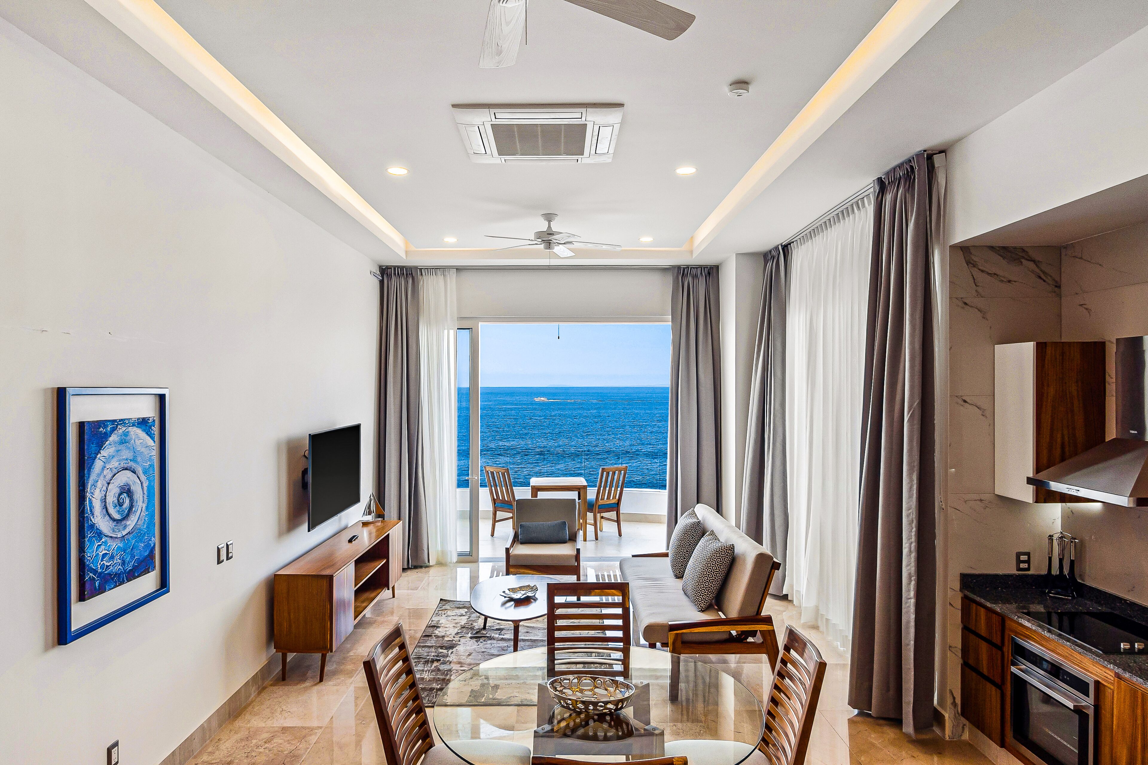 A modern living room with a large window overlooking the sea, a ceiling fan, and a flat-screen TV.