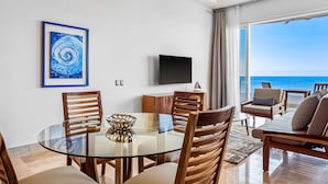 A modern dining area with a glass table, wooden chairs, a TV on the wall, and a view of the ocean. - Blue Horizon 401 | 1BR/2BA | PV Ocean View, Pool (Puerto Vallarta)