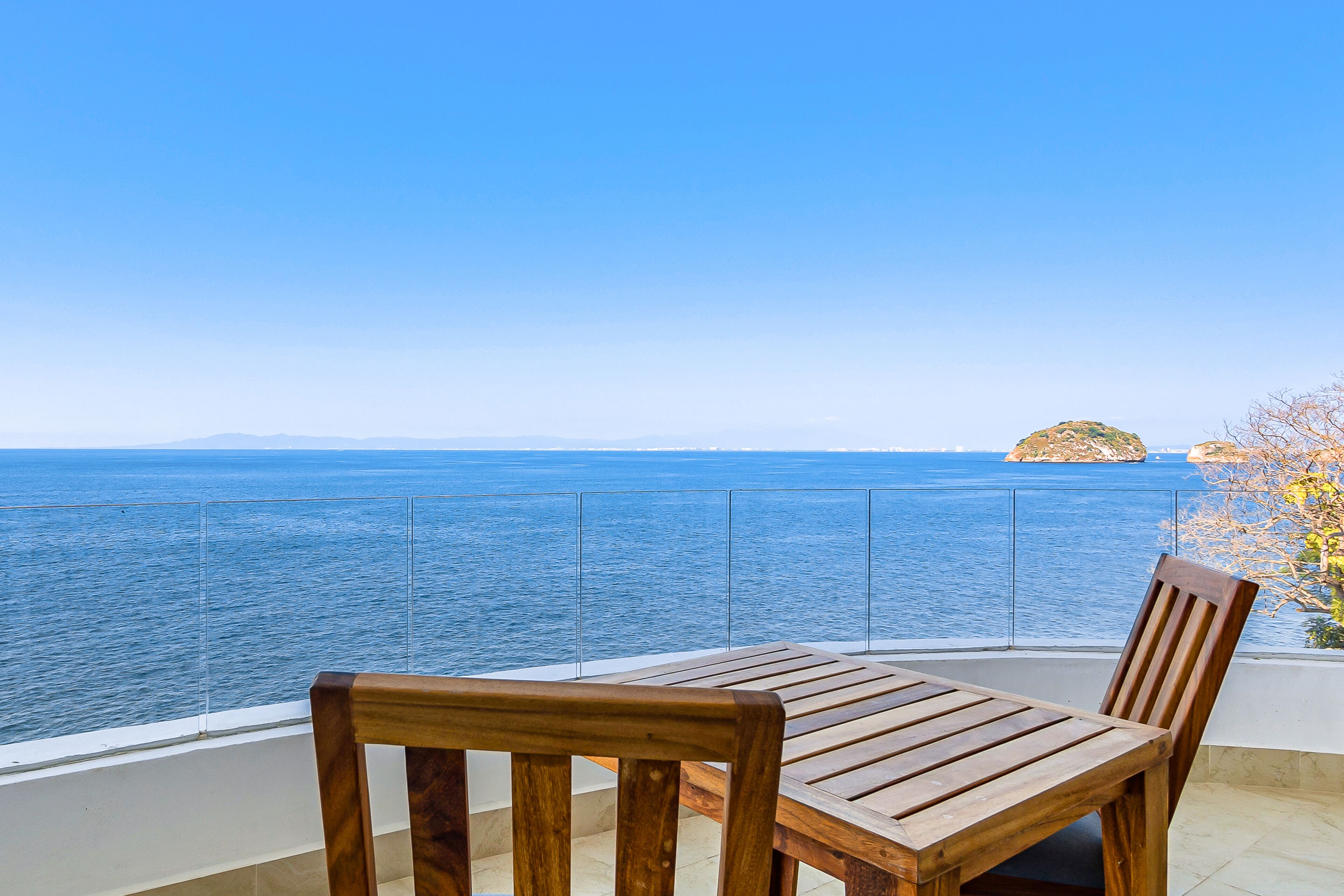 A wooden table and chair on a balcony with a clear view of the sea and a distant island.