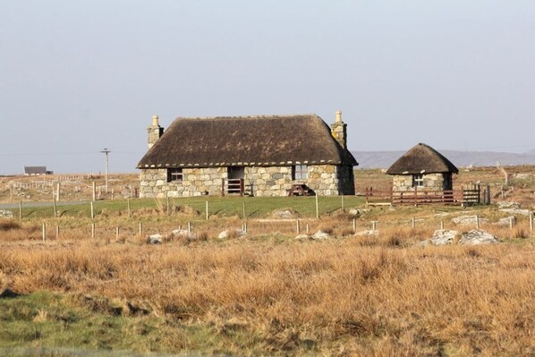 A stone house with a thatched roof, a fenced yard, and a rural landscape.