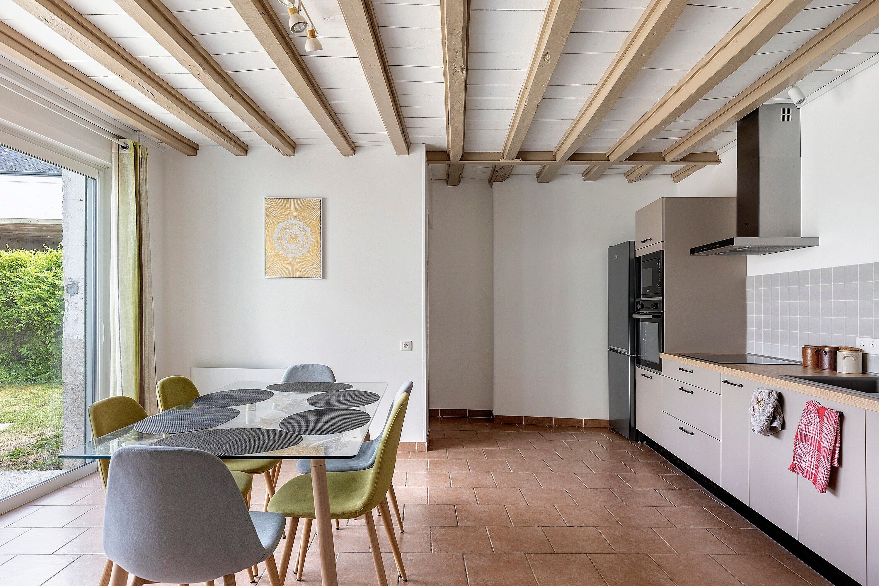 A modern kitchen with a dining area, wooden ceiling beams, and a view of greenery outside.