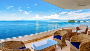 Ein Balkon mit Korbmöbeln, einem Glastisch und Blick aufs Meer.