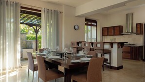 A dining area with a long table set for six, a kitchen with wooden cabinets, and a clock on the wall. - Peaceful 4-Bedroom Villa / Infinity Pool & Garden View (Bel Ombre)