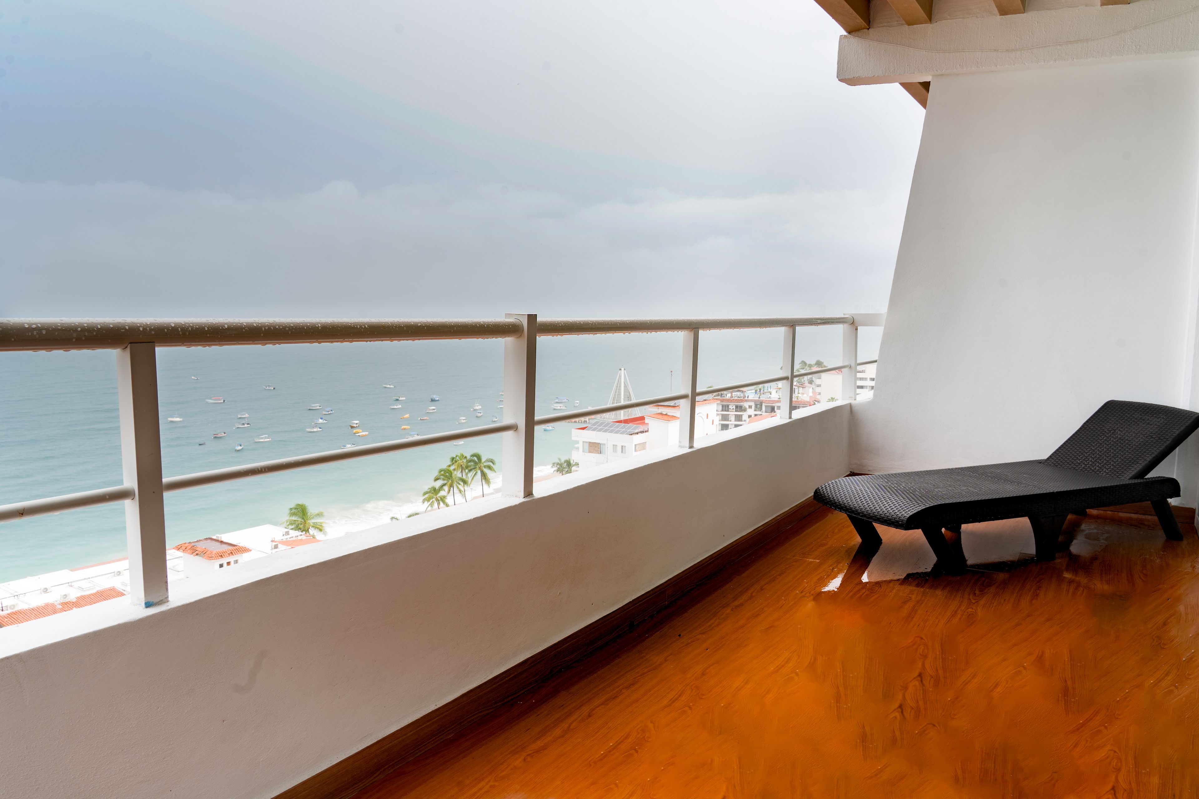 Un balcón con vista al mar y la playa, una silla de descanso negra y suelo de madera.