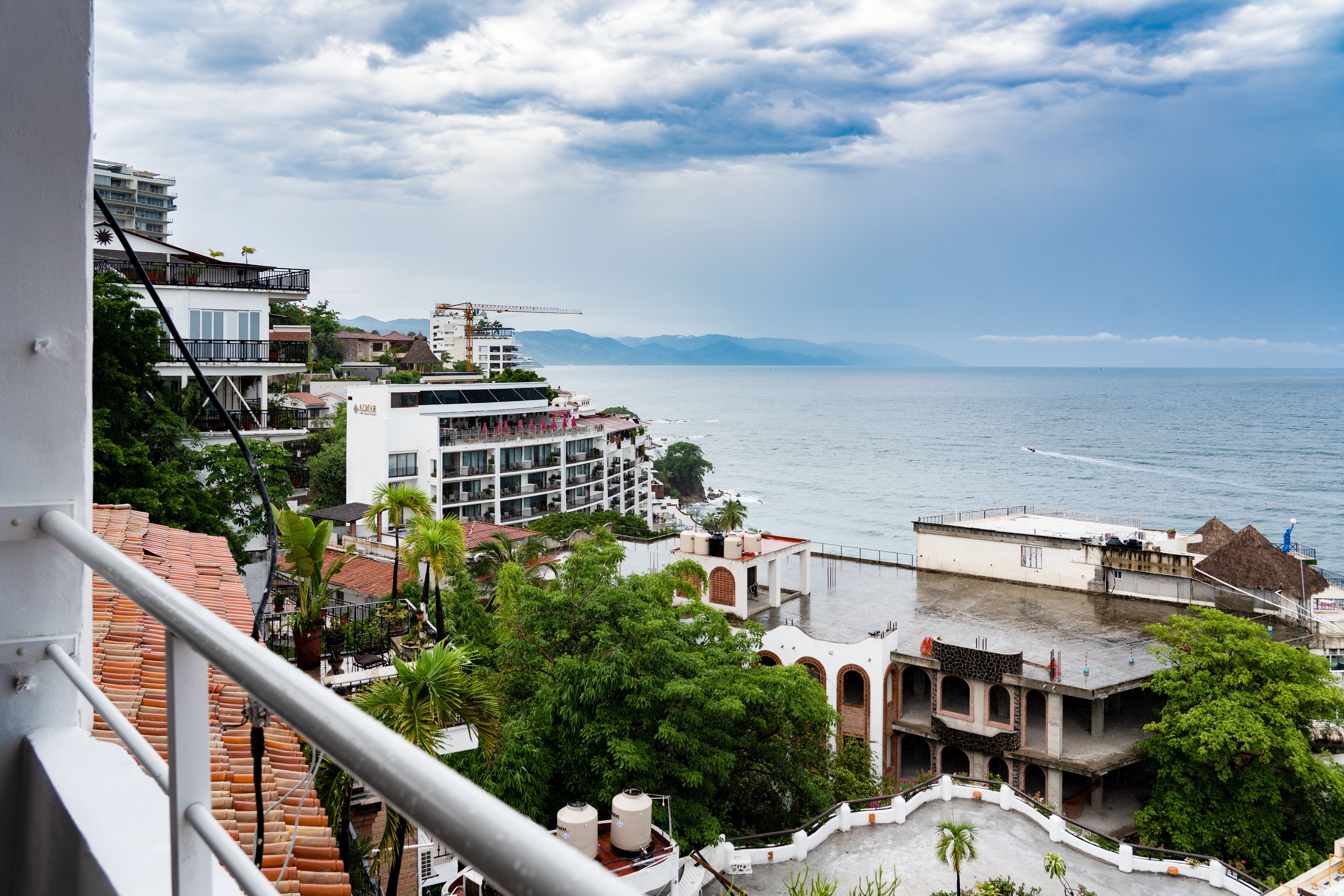 Un hôtel en bord de mer doté de nombreux balcons donnant sur la mer, une grue de construction en arrière-plan et une vue sur des montagnes lointaines.