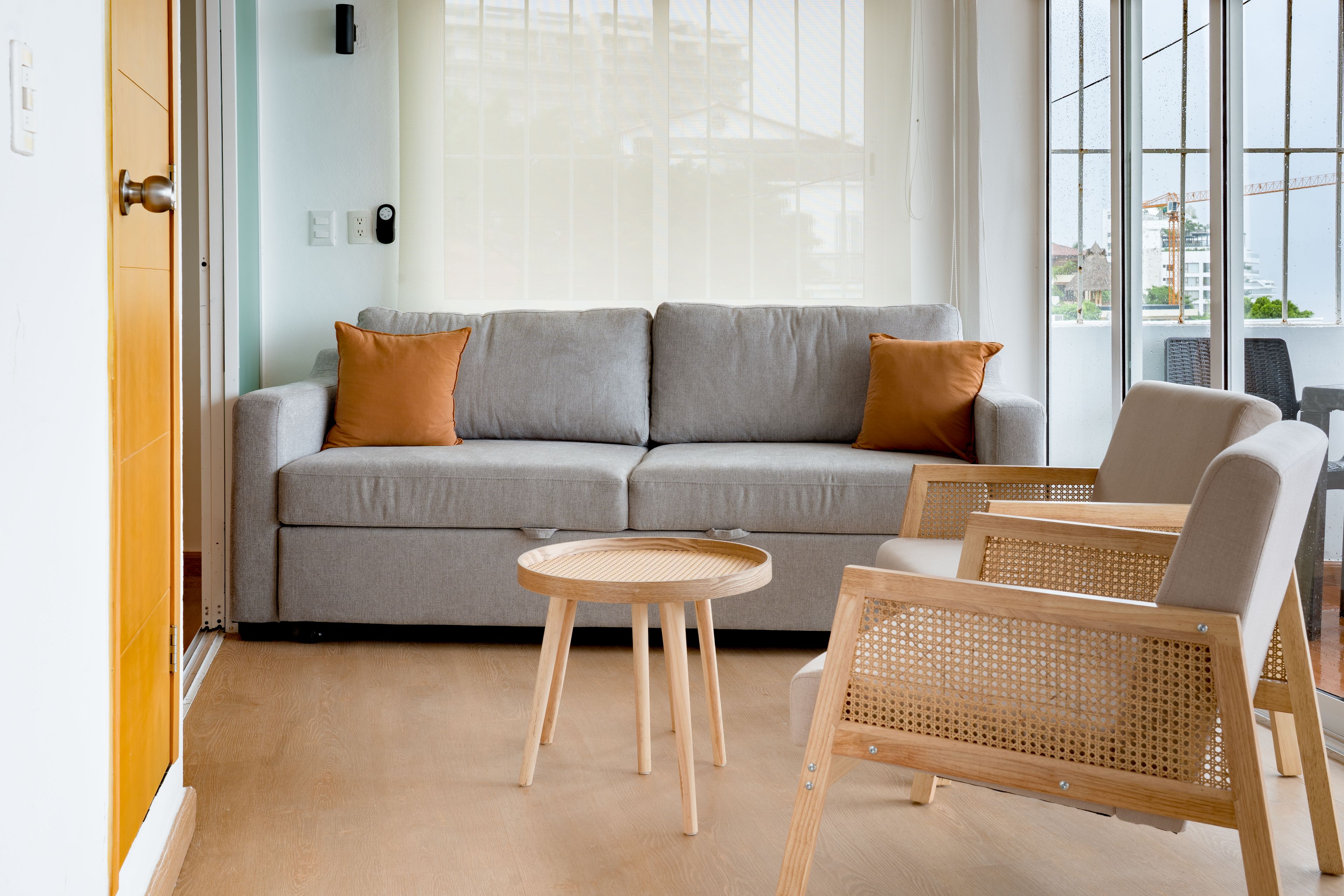 A modern living room with a grey sofa, two brown cushions, a round wooden side table, and two wicker chairs.