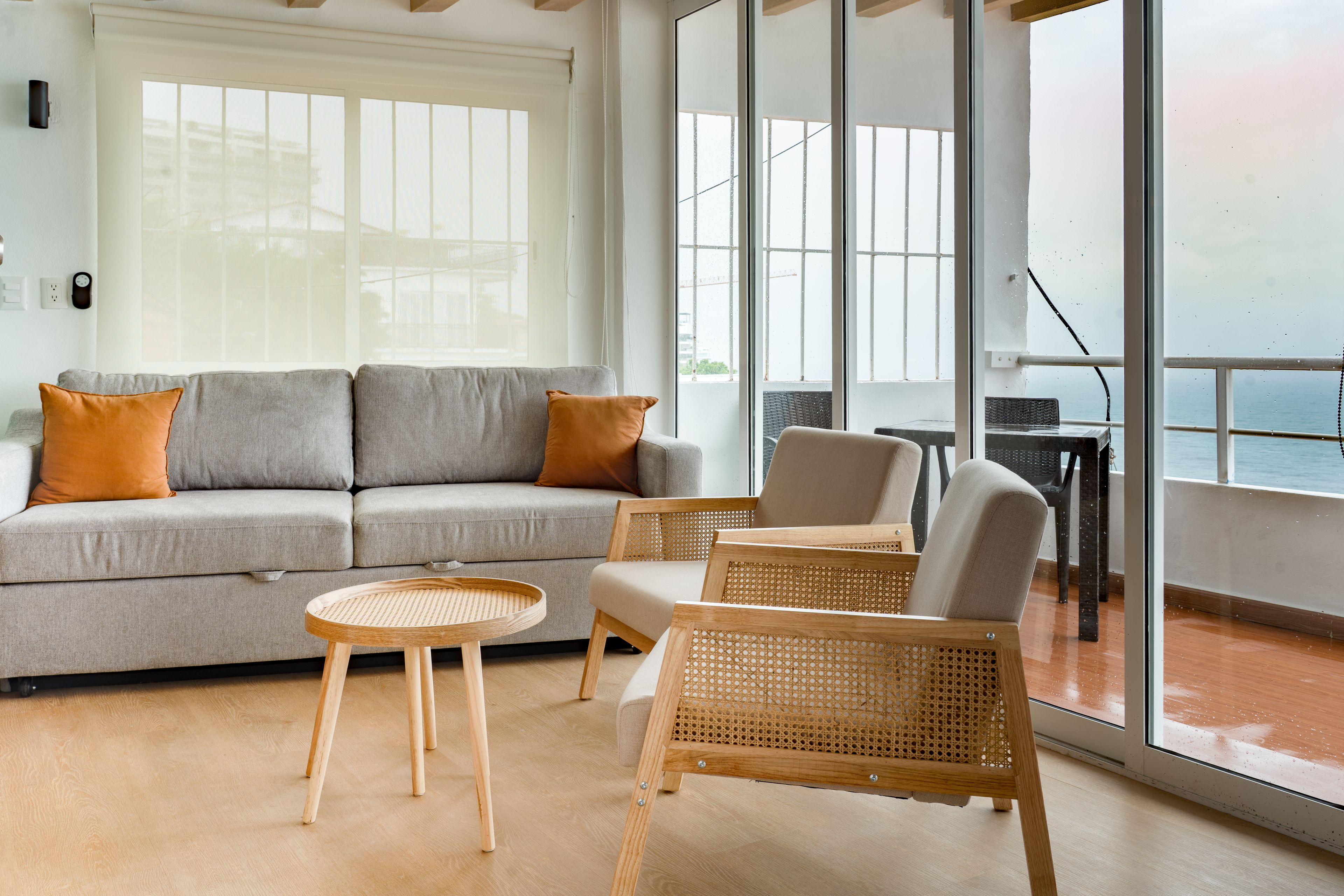 Un salon moderne avec un canapé gris, deux fauteuils, une petite table ronde et une vue sur la mer grâce à de grandes fenêtres.