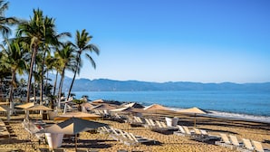 Ein Strand mit Reihen von Liegestühlen und Sonnenschirmen, Palmen und Blick auf das Meer und die Berge.