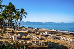 A beach with rows of sun loungers and umbrellas, palm trees, and a view of the ocean and mountains. - Puerto Vallarta 3BR Ocean View Condo w/ Pool & Gym (Puerto Vallarta)